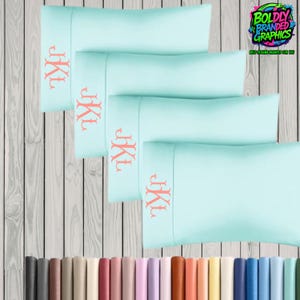 May include: Four light blue pillowcases with coral-colored monogrammed initials. The pillowcases are stacked on top of each other, with the initials visible on the top of each pillowcase. The image also includes a logo that says "Boldly Branded Graphics".