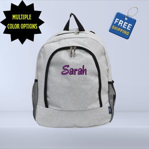 May include: A silver glitter backpack with black trim and mesh side pockets. The name "Sarah" is embroidered in purple on the front. The backpack has a top handle and two zippered compartments. The image also includes text that says "Multiple Color Options".