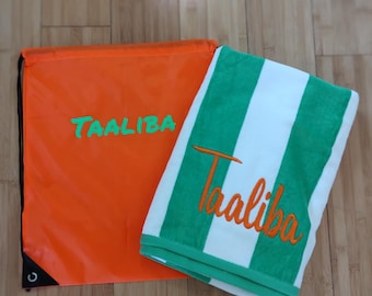 Puede incluir: Una bolsa naranja con cordón y una toalla a rayas verdes y blancas. Ambos artículos tienen el nombre "TAALIBA" bordado en un color contrastante. La toalla tiene rayas verdes y un borde verde.