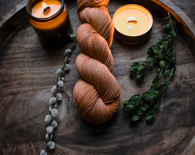 May include: A skein of orange yarn, two lit candles, and a sprig of green foliage on a wooden tray.