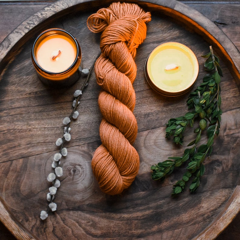 May include: A wooden tray with two lit candles, a sprig of greenery, and a ball of orange yarn.