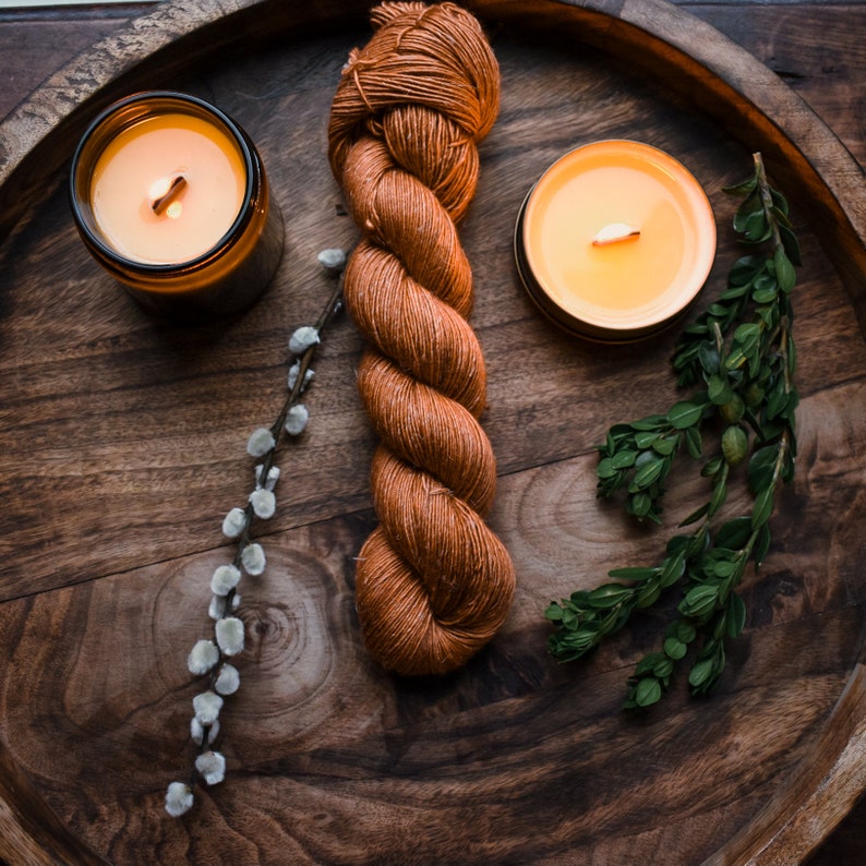 May include: A wooden tray with a ball of orange yarn, two lit candles, and a sprig of green foliage.