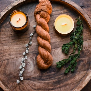 May include: A wooden tray with two lit candles, a skein of orange yarn, a sprig of green leaves, and a sprig of white pussy willow.