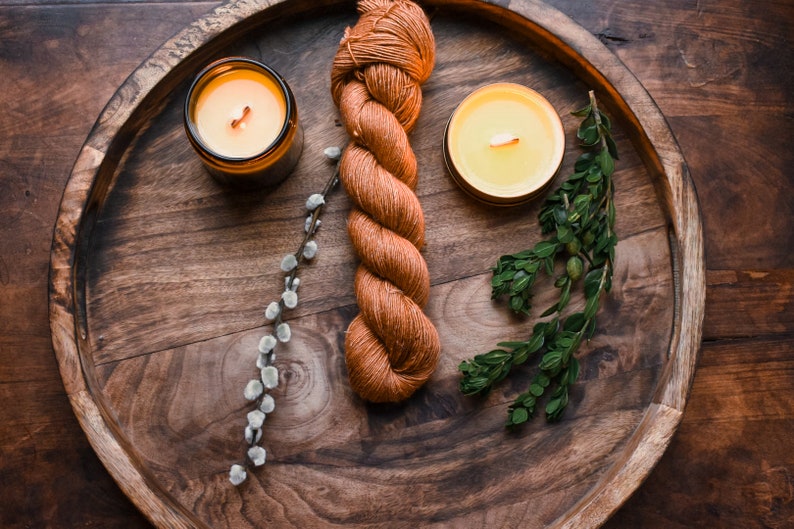 May include: A wooden tray with two lit candles, a skein of orange yarn, a sprig of green leaves, and a sprig of white pussy willow.