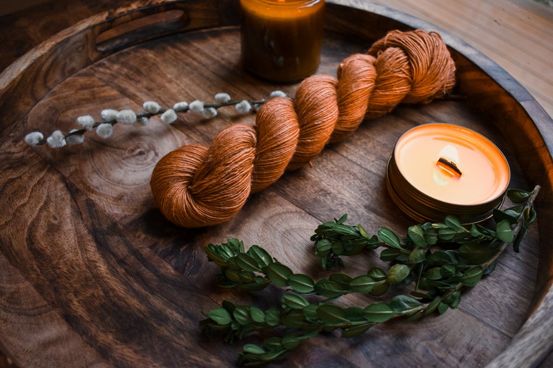 May include: A wooden tray with a skein of orange yarn, a lit candle, and sprigs of greenery.