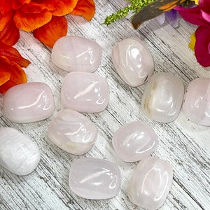 May include: A collection of smooth, polished rose quartz stones in varying sizes. The stones are a soft pink color and have a slightly translucent appearance. The background features a wooden surface and colorful flowers.