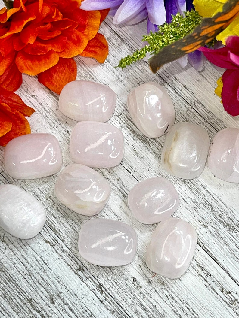 May include: A collection of smooth, polished rose quartz stones in varying sizes. The stones are a soft pink color and have a slightly translucent appearance. The background features a wooden surface and colorful flowers.