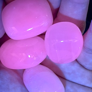 May include: Four smooth, rounded, pink stones are held in a hand. The stones have a glossy finish and vary slightly in shape. The lighting gives them a soft, glowing appearance, highlighting their polished surfaces. These stones could be used for decoration or as a craft supply.
