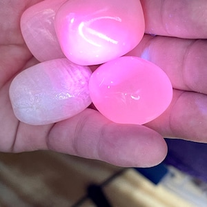 May include: Four smooth, polished pink stones are held in a hand. The stones vary in shade from pale to vibrant pink, with a glossy, reflective surface. The stones are likely gemstones or decorative rocks.