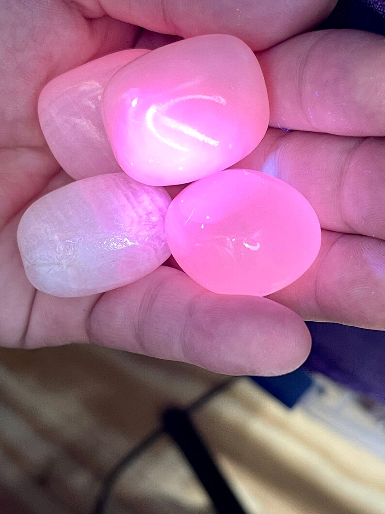 May include: Four smooth, polished pink stones are held in a hand. The stones vary in shade from pale to vibrant pink, with a glossy, reflective surface. The stones are likely gemstones or decorative rocks.