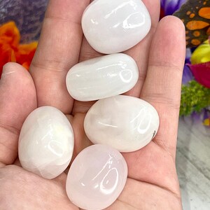 May include: Five smooth, polished stones in varying shades of white and pink are held in a hand. The stones have an oval shape and a glossy finish. The background includes hints of orange and purple flowers.
