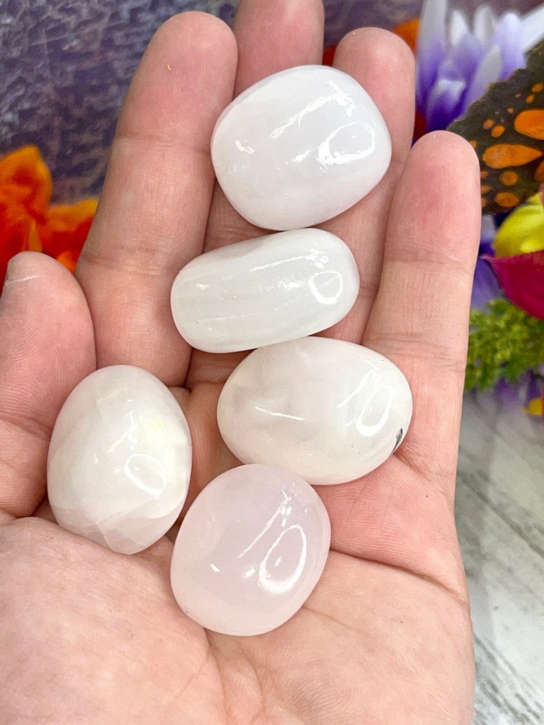 May include: Five smooth, polished stones in varying shades of white and pink are held in a hand. The stones have an oval shape and a glossy finish. The background includes hints of orange and purple flowers.