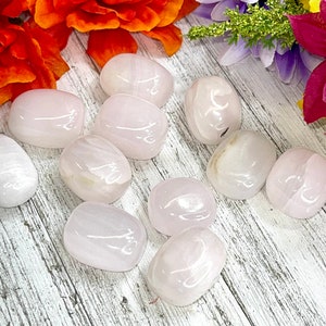 May include: A collection of smooth, polished rose quartz stones. The stones are a soft pink color with subtle variations in tone. They are arranged on a white and gray wooden surface, with vibrant orange and purple flowers in the background.