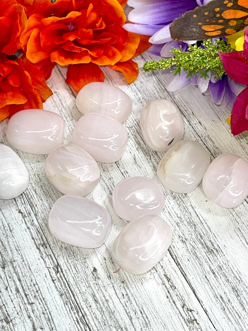 May include: A collection of smooth, polished rose quartz stones. The stones are a soft pink color with subtle variations in tone. They are arranged on a white and gray wooden surface, with vibrant orange and purple flowers in the background.