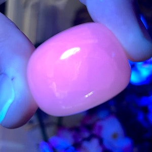 May include: A smooth, rounded, pink gemstone is held between two fingers. The stone has a glossy surface and reflects light, appearing to glow. The background is blurred, with hints of blue and purple.
