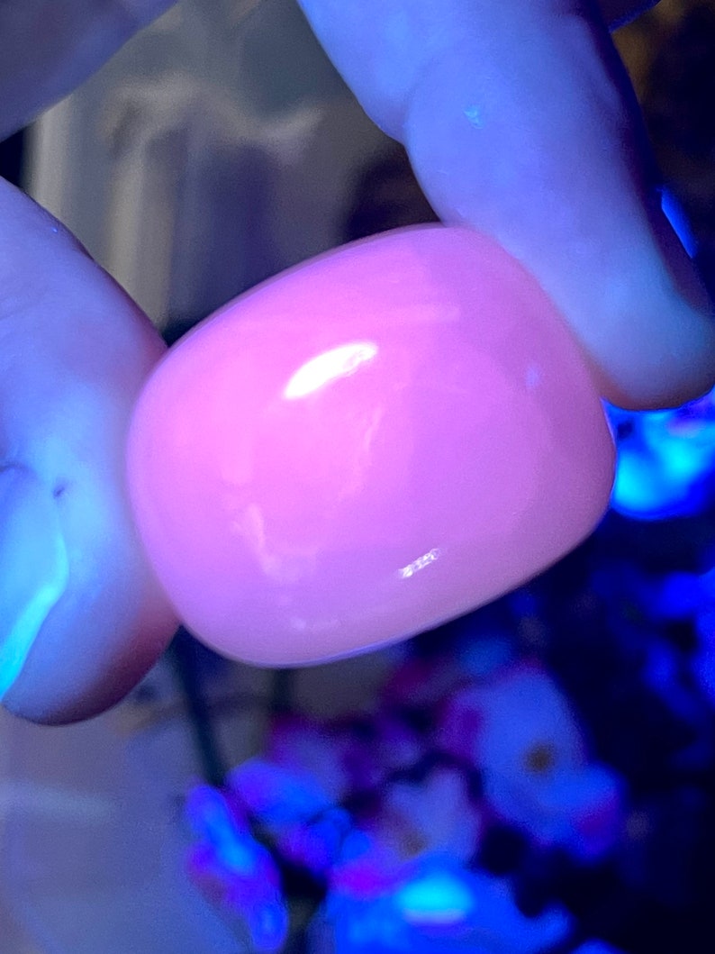 May include: A smooth, rounded, pink gemstone is held between two fingers. The stone has a glossy surface and reflects light, appearing to glow. The background is blurred, with hints of blue and purple.