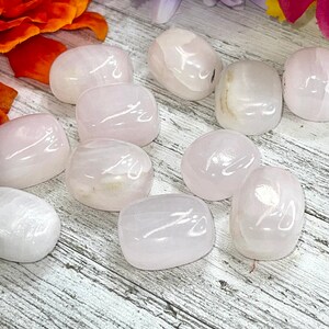 May include: A collection of smooth, polished rose quartz stones. The stones are oval-shaped and vary in shades of pale pink. The background features a wooden surface and colorful flowers, enhancing the natural beauty of the crystals.