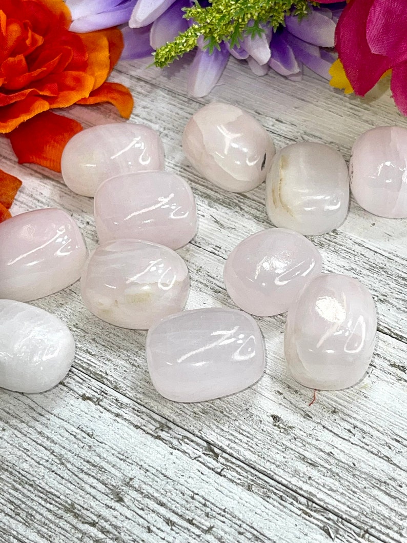 May include: A collection of smooth, polished rose quartz stones. The stones are oval-shaped and vary in shades of pale pink. The background features a wooden surface and colorful flowers, enhancing the natural beauty of the crystals.