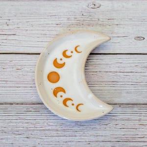May include: A white ceramic crescent moon dish, decorated with orange moon phase symbols. The dish is displayed on a light-coloured wooden surface. This decorative dish is likely used for holding small items, such as jewellery or trinkets.