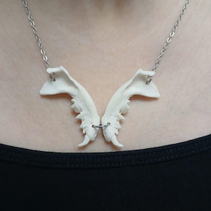 May include: A unique necklace featuring a pendant shaped like a butterfly, crafted from off-white bone-like material. The pendant is suspended from a delicate silver-colored chain. The necklace is worn against a black top.