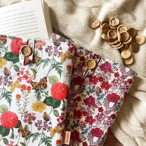 Puede incluir: Dos cubiertas de libros con patrones florales. Una cubierta es blanca con flores rojas y mariposas, la otra es crema con flores rojas y burdeos. Ambas cubiertas tienen un botón de madera y el texto "bunny books" impreso en la parte inferior.