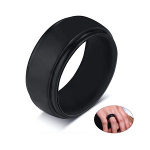May include: A solid black ring with a flat top and a grooved edge. The ring is shown in a close-up view, with a smaller inset image of the ring on a finger. The ring is made of a matte material.