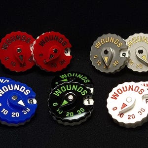 May include: Six different coloured wound trackers for role-playing games. The trackers are circular and have a dial with numbers 0, 10, 20, and 30. The word "WOUNDS" is printed on each tracker. The trackers are red, blue, black, white, and two shades of grey.
