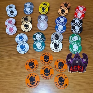 May include: A collection of colorful game tokens with a skull graphic and the words "Battle Shocked." The tokens are in various colors, including white, blue, green, orange, and red. A sticker with a purple Grim Reaper is also present.