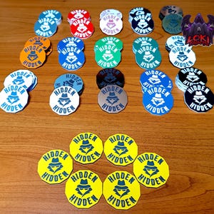 May include: A collection of circular stickers in various colours including yellow, orange, red, blue, green, black, and white. Each sticker features a graphic of a person wearing a fedora hat and the text HIDDEN printed above and below the graphic.