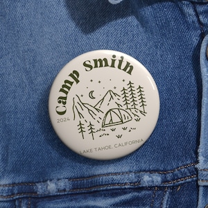 May include: A white button with a green outline and the text "Camp Smith 2024" and "Lake Tahoe, California". The button features a line drawing of a mountain range, a crescent moon, stars, and a tent.
