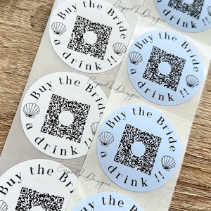 May include: Sheet of round stickers with the text "Buy the Bride a drink !!" in black, with a QR code in the center and seashell graphics. Some stickers are white, others are light blue.