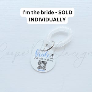May include: A white round tag with the text "I'm the bride buy me a drink!" in blue and a ring icon. The tag is attached to a white plastic hair scrunchie. The text "I'm the bride - SOLD INDIVIDUALLY" is written in black at the top of the image.