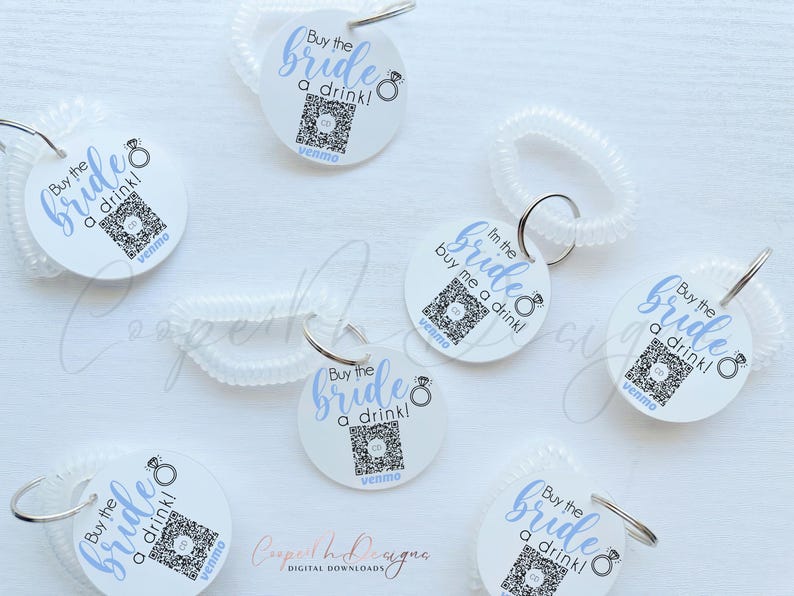 May include: Six white circular tags with blue text that reads "Buy the bride a drink!" and a blue ring icon. Each tag has a QR code and the word "venmo" printed on it. The tags are attached to clear plastic coil keychains.