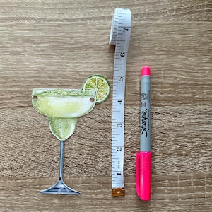 May include: A green and yellow margarita glass with a lime wedge on the rim. A pink Sharpie marker and a white measuring tape are next to the glass.