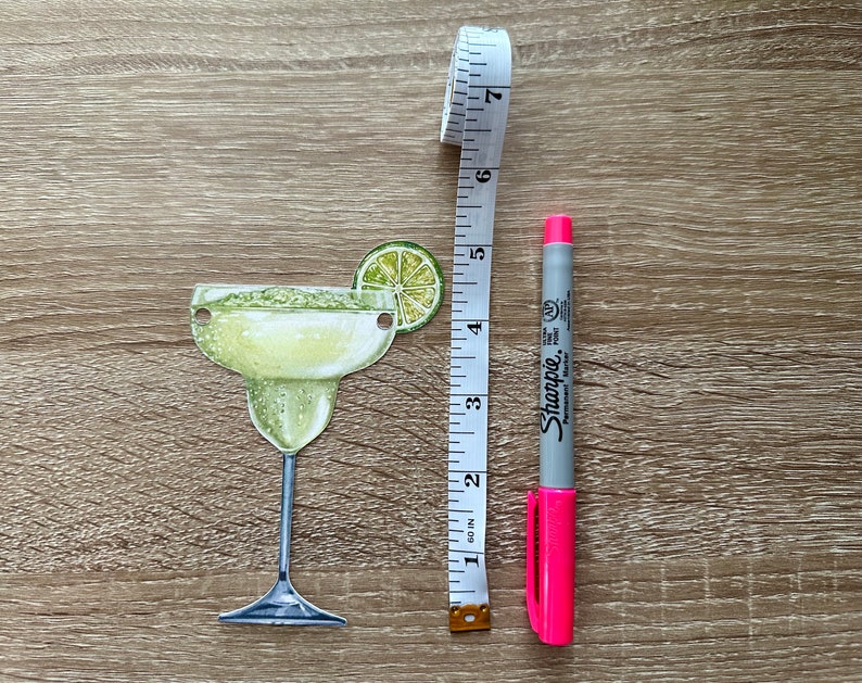 May include: A green and yellow margarita glass with a lime wedge on the rim. A pink Sharpie marker and a white measuring tape are next to the glass.