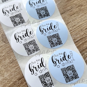 May include: A roll of round stickers with the text "Buy the Bride a drink!" in black script. Each sticker has a QR code and a champagne glass illustration. Some stickers are light blue, others are white.
