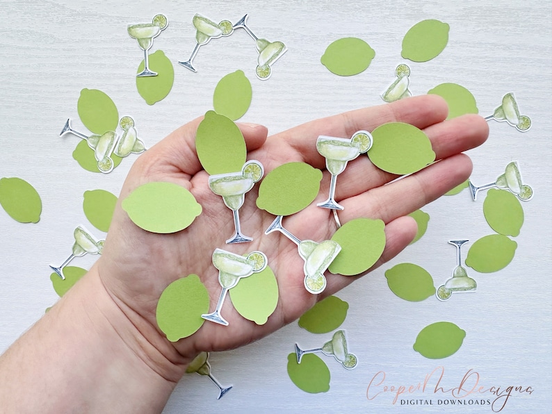 May include: A hand holding a variety of green lime and margarita glass confetti. The confetti is made of paper and features a lime wedge and a margarita glass with a lime wedge garnish.