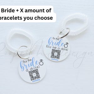 May include: Two white circular tags with blue text and a ring design. The text on the tags reads "Buy the bride a drink!" and "I'm the bride buy me a drink!" Each tag has a QR code and the word "venmo" printed below. The tags are attached to clear plastic bracelets.