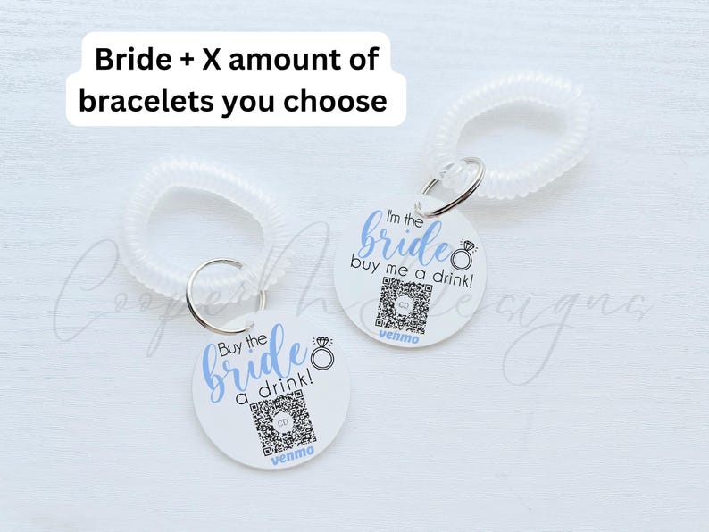 May include: Two white circular tags with blue text and a ring design. The text on the tags reads "Buy the bride a drink!" and "I'm the bride buy me a drink!" Each tag has a QR code and the word "venmo" printed below. The tags are attached to clear plastic bracelets.