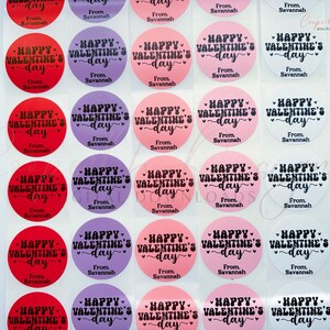 Puede incluir: Una hoja de 24 pegatinas redondas con tem&aacute;tica de San Valent&iacute;n. Las pegatinas son de tres colores: rojo, rosa y morado. Cada pegatina tiene un fondo blanco con texto negro que dice "Happy Valentine's Day" con un dise&ntilde;o de coraz&oacute;n. El texto "From Savannah" est&aacute; impreso debajo del texto principal en cada pegatina.
