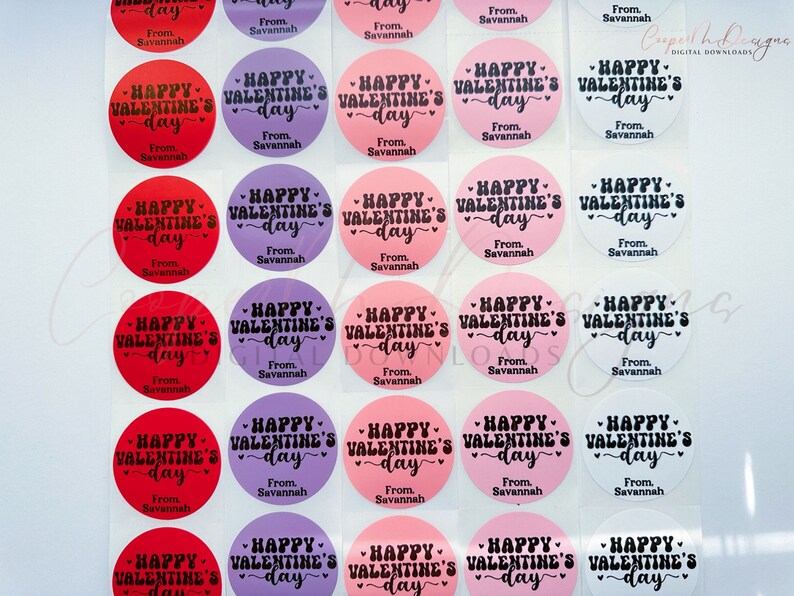 Puede incluir: Una hoja de 24 pegatinas redondas con tem&aacute;tica de San Valent&iacute;n. Las pegatinas son de tres colores: rojo, rosa y morado. Cada pegatina tiene un fondo blanco con texto negro que dice "Happy Valentine's Day" con un dise&ntilde;o de coraz&oacute;n. El texto "From Savannah" est&aacute; impreso debajo del texto principal en cada pegatina.