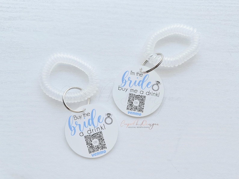 May include: Two white circular tags with black text and a blue ring design. The text on the tags reads "Buy the bride a drink!" and "I'm the bride buy me a drink!" with the Venmo logo at the bottom of each tag.