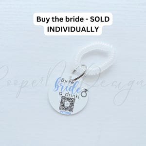 May include: White round keychain with a blue and black design that says "Buy the bride a drink!" with a ring icon and a Venmo logo. The keychain is attached to a clear plastic coil.
