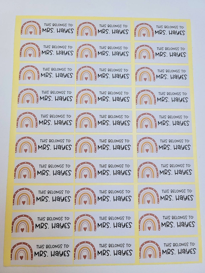 May include: A sheet of 18 personalized name labels with a rainbow design. Each label features a rainbow with a heart in the center and the text "THIS BELONGS TO MRS. HAYES".