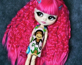 Könnte beinhalten: Eine Puppe mit pinken, lockigen Haaren, die ein weißes Kleid mit einem Affenmuster und blauen und grünen Akzenten trägt. Die Puppe trägt blaue Socken und weiße Schuhe.