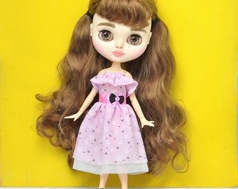 Könnte beinhalten: Eine Puppe mit langen braunen Haaren, die ein rosa Kleid mit weißen Blumen und einer rosa Schleife trägt. Die Puppe trägt schwarze Schuhe und hat braune Augen.