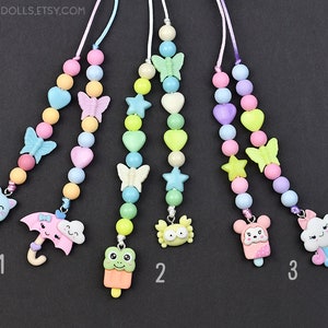 Peut inclure: Trois colliers de perles colorés avec différents pendentifs. Le premier collier a un pendentif en forme de cornet de glace avec un chat, le deuxième a un pendentif en forme de cornet de glace avec une grenouille et le troisième a un pendentif en forme de cornet de glace avec un ours.