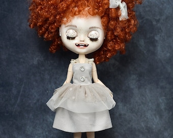Könnte beinhalten: Eine Puppe mit roten Locken, die ein weißes Kleid mit einem mehrschichtigen Rock und weißen Knöpfen trägt. Die Puppe hat aufgemalte Wimpern und ein zahniges Lächeln.