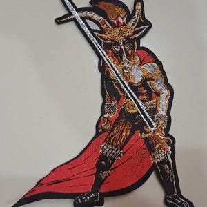 May include: Embroidered patch depicting a horned figure in a red cape, wielding a sword. The figure has a goat-like face and is rendered in black, red, gold, and silver. Suitable for application on clothing or accessories.