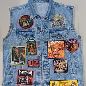 以下が含まれることがあります： Slayer、Motörhead、Manowar、W.A.S.P.、Mercyful Fateなどの音楽バンドの刺繍パッチが施されたライトブルーのデニムベスト。この袖なしジャケットは、ボタン留めのフロント、2つの胸ポケット、クラシックな襟のデザインが特徴です。